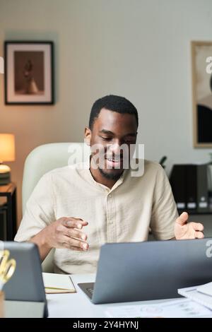 Smiling man sitting at desk in office setting, using laptop. Engaging in conversation while holding gesturing hand in relaxed office environment Stock Photo