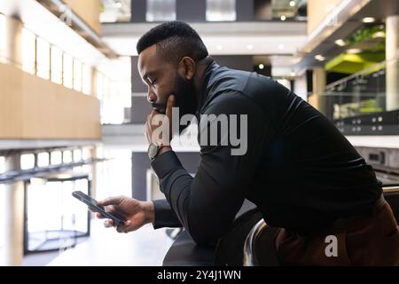 Leaning on railing, african american businessman using smartphone and thinking in modern office Stock Photo