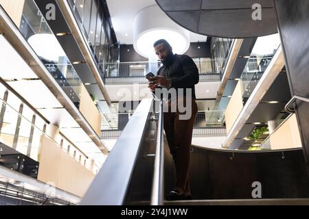 Businessman using smartphone while walking down stairs in modern office building, copy space Stock Photo