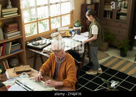 Elderly woman typing on computer in home office with plants and vinyl records while younger woman writes notes nearby Stock Photo