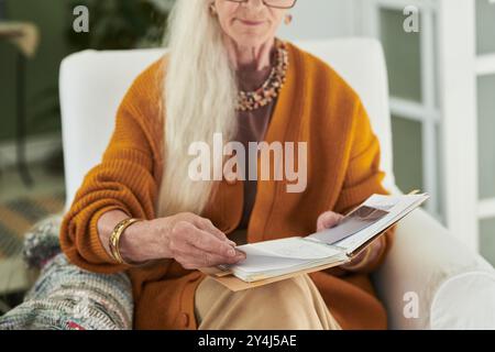 Elderly woman with long gray hair reading book while sitting comfortably in chair at home, showing peaceful and relaxed moment Stock Photo