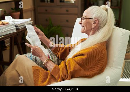 Elderly woman with glasses smiling while reading a letter, wearing an orange sweater and sitting in a cozy, well-decorated living room with plants and Stock Photo
