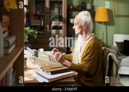 Elderly woman working on typewriter in cozy home office with bookshelves and green walls surrounding her. She holds a pair of glasses while concentrating on her work Stock Photo