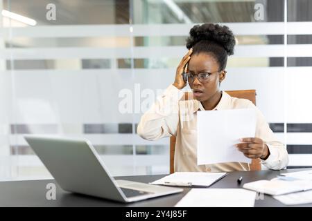 Young African American woman at workplace feeling stressed while working in office. Holding documents, looking at laptop with hand on forehead, experiencing workplace stress, frustration during work Stock Photo