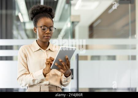 Young african american woman using tablet at workplace. Engaged in work and demonstrating productivity in modern office setting. Professional attire and focus on task. Stock Photo