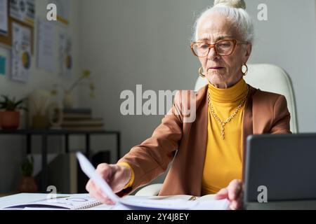Elderly woman sitting at desk working with documents in modern office environment. Wearing glasses and focused on task, business charts and office supplies visible in background Stock Photo