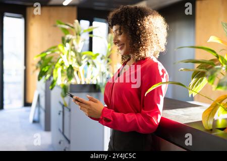 Using smartphone, woman in red blouse standing in modern office environment Stock Photo
