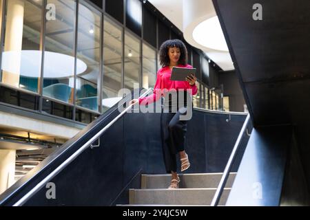 Businesswoman walking down stairs using tablet in modern office building Stock Photo