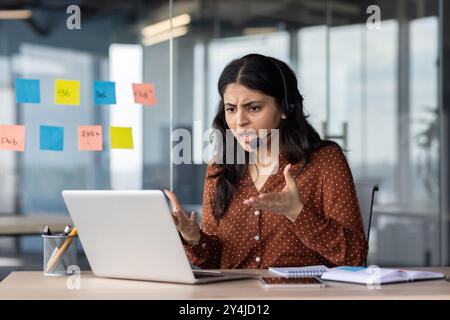 Angry woman yelling on online video call, support worker with headset working inside office using laptop, frustrated with interlocutor. Stock Photo