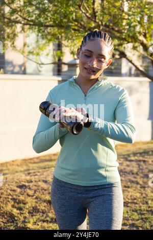 Checking smartwatch, woman holding water bottle, exercising outdoors in sunlight Stock Photo