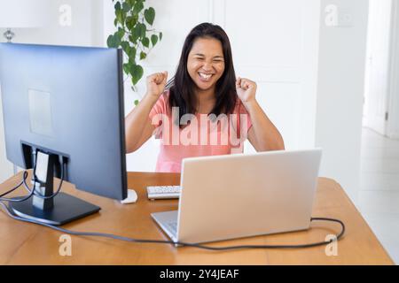 Celebrating success, woman with curly hair using laptop and desktop computer at home office Stock Photo