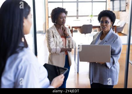 Discussing project, businesswomen using laptop and tablet in modern office setting Stock Photo