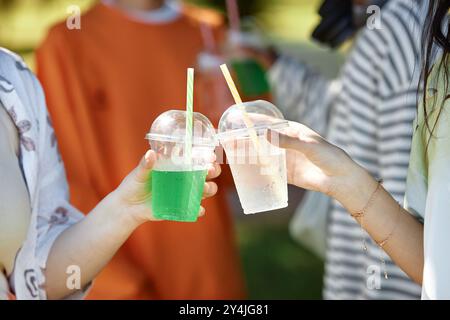 Close up on hands of two teenage girls holding plastic takeaway cups with refreshing drinks clinking glasses while hanging out in park enjoying summer break, copy space Stock Photo