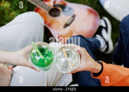 Close up on hands of teenagers holding plastic cups with refreshing drinks while clinking glasses and enjoying picnic in park on sunny summer day, copy space Stock Photo