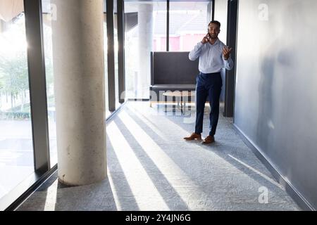 Talking on smartphone, businessman in formal attire walking in modern office hallway, copy space Stock Photo