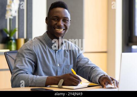 Smiling african american businessman writing in notebook at desk with laptop and smartphone Stock Photo