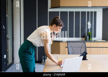 Talking on smartphone, woman taking notes while working on laptop in office Stock Photo
