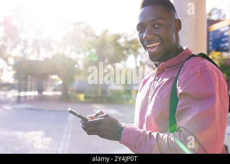 Smiling man using smartphone outdoors, wearing backpack, enjoying sunny day, copy space Stock Photo