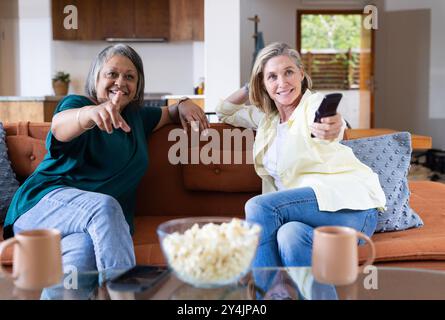 At home, watching tv and eating popcorn, senior multiracial female friends relaxing on couch Stock Photo