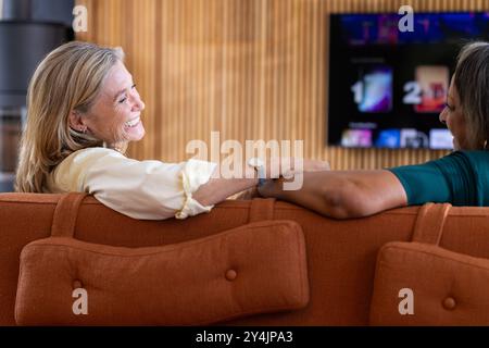 At home, smiling senior multiracial female friends sitting on couch, watching tv and conversation Stock Photo