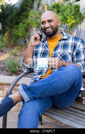 Talking on smartphone, man sitting on bench holding coffee cup, smiling outdoors Stock Photo