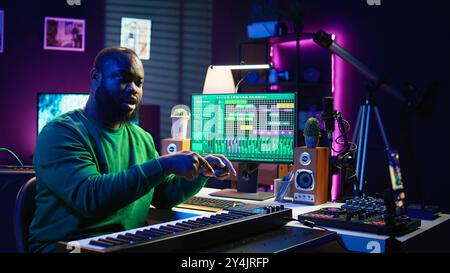 Musician doing online paid classes to help people play piano notes, teaching lessons from his home studio and recording video tutorials. Musical instructor playing midi controller keyboard. Camera B. Stock Photo