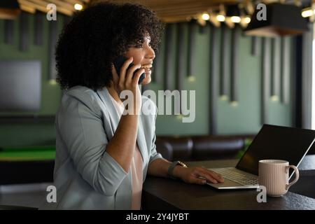 Talking on smartphone, woman using laptop and drinking coffee in modern office Stock Photo