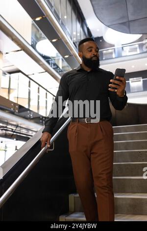 Walking down stairs, African american businessman using smartphone in modern office building Stock Photo