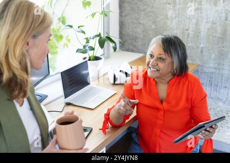 Senior multiracial female colleagues using tablet and laptop while having coffee in office Stock Photo
