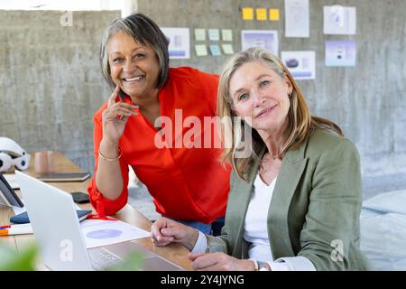 Smiling senior multiracial female colleagues working together on laptop in modern office setting Stock Photo