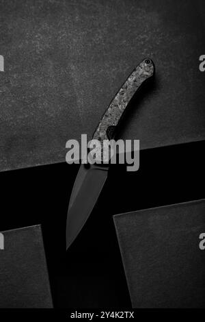 Minimalistic photo of a folding knife on a dark, textured surface. The sleek black blade and handle design create a modern, tactical look, ideal for o Stock Photo