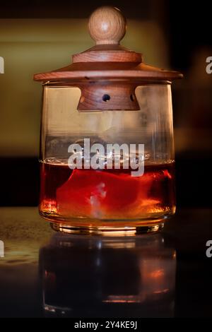 A smoked whiskey cocktail in a glass with a wooden cover. The rich amber liquid is illuminated against a dark, background, subtle smoke visible inside. Stock Photo