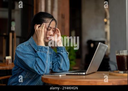 A side view of a stressed, thoughtful Asian woman in a denim jacket, feeling headache and tired, facing a problem with her project while working on he Stock Photo