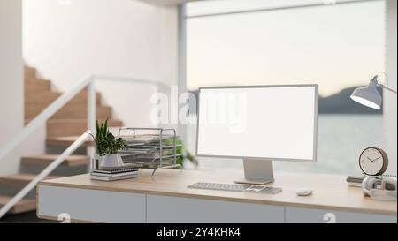 A computer desk in a bright, contemporary home, featuring a large glass window behind it, with stairs nearby and a computer displaying a white screen Stock Photo