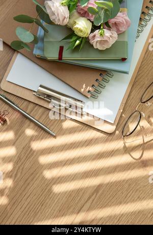 Bright sunlight filters through the blinds, casting shadows on a wooden table adorned with colorful notebooks, fresh flowers, and stylish stationery, Stock Photo