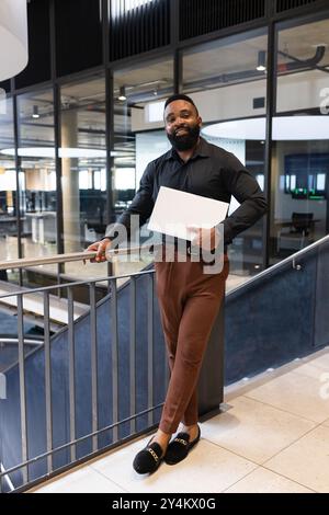 Holding laptop, African American businessman standing confidently in modern office building lobby Stock Photo