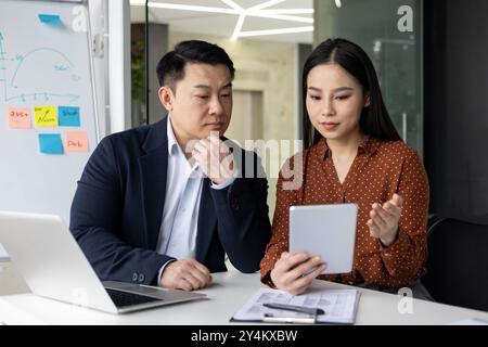Asian business professionals engaging in collaborative discussion using tablet and laptop. Man and woman focused on productive teamwork and planning at modern office workspace. Stock Photo