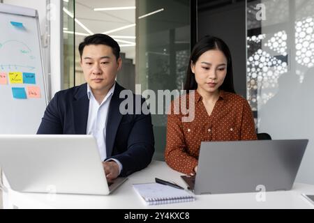 Asian business professionals working on laptops in contemporary office. Collaboration and focus on digital tasks, demonstrating teamwork and productivity. Stock Photo