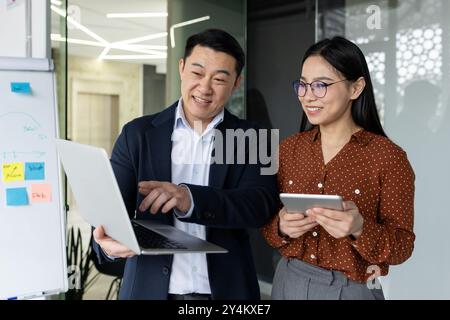 Asian business team discussing strategies using laptop and tablet. Collaborative work environment, showcasing teamwork communication between colleagues. Professionals engaging in productive meeting Stock Photo