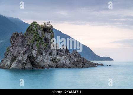 At dusk. Gentle waves lap against smooth, rounded rocks in a tranquil cove. The sky is painted in soft hues of pink and purple, reflecting on the calm Stock Photo