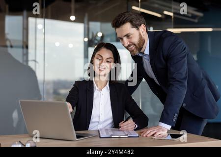 Business professionals engaging in teamwork at office. Smiling colleagues collaborating on laptop, analyzing data and discussing strategies. Professional attire and modern office environment Stock Photo