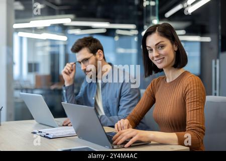 Two business professionals collaborating in a modern office environment. Woman smiling while typing on laptop, man in background working on laptop with thoughtful expression Stock Photo