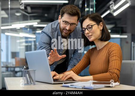 Two colleagues collaborating in office, focused on project. Man pointing at laptop screen, woman typing. Teamwork, technology, and planning concepts. Pair engaged with office work Stock Photo