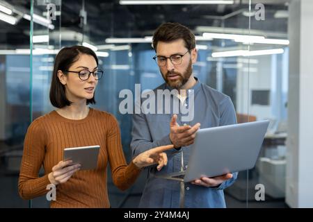 Colleagues engage in discussion in modern office setting. Woman holds tablet, man gestures at laptop while actively collaborating. Bright, open workspace enhances teamwork and productivity Stock Photo