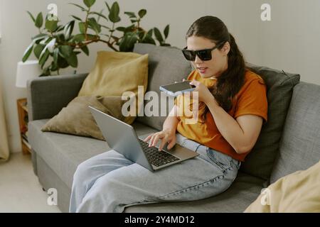 Young brunette woman with disability speaking on smartphone while using laptop in living room Stock Photo