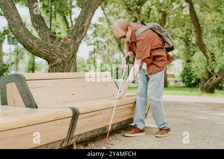 Caucasian blind man using metal cane to walk in park alone, he touching bench Stock Photo