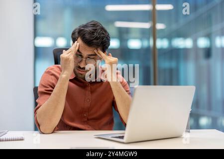 Businessman experiencing stress and exhaustion at indoor workplace, holding head with both hands. Office setting includes laptop reflecting pressure and frustration in professional environment. Stock Photo