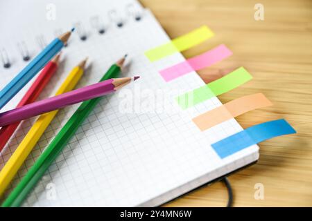 Colored pencils on a part of a spiral notebook with squared paper and narrow sticky notes on a wooden desk, office or school supplies, copy space Stock Photo