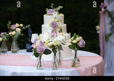 White three-tiered wedding cake decorated with roses and eustoma flowers standing on a table with a pink tablecloth outdoors Stock Photo