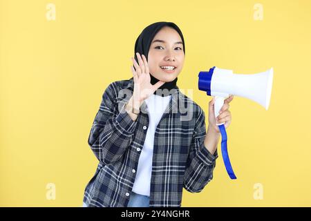 Young Asian Indonesian Muslim woman in Headscarf  Veil Hijab Speaking Through Megaphone, Isolated on Yellow Background Stock Photo